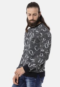 Grijze sweater met een opvallende witte letterprint, ronde halslijn en zwarte ribgebreide manchetten en zoom. Draag met blauwe versleten spijkerbroek.