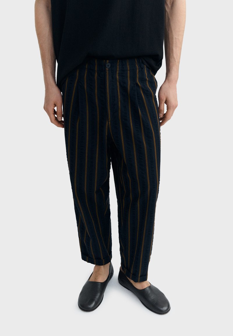 Personne portant un pantalon noir ample avec des fines rayures verticales orange et des chaussures noires sans lacets, sur un fond clair uni.
