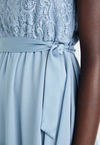 Svetlo modra obleka z čipkastim bodicem in mehkim krilom. Tkaninski pas, zavezan v pasu, doda podrobnost. Gladka tekstura in lahkotna zasnova.