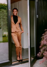 Brauner maßgeschneiderter Anzug mit einer eng anliegenden Jacke und schmal zulaufenden Hosen, kombiniert mit einem schwarzen Rollkragenpullover und schwarzen High Heels, steht in einer Tür.