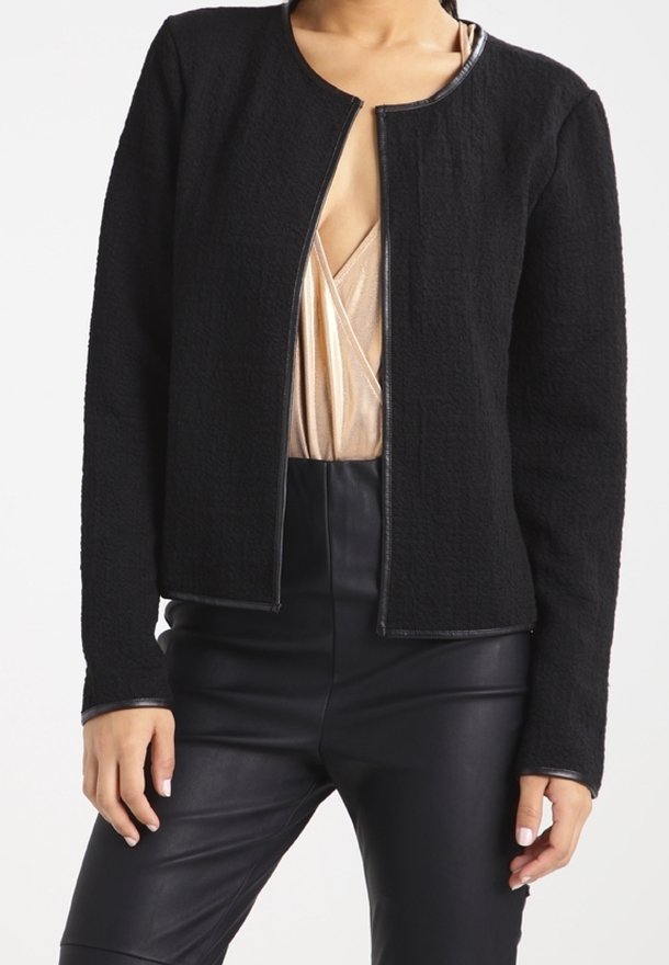 Femme portant une veste texturée noire ouverte sur un haut beige à col en V et un pantalon en cuir noir taille haute.