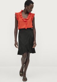 Rote Rüschbluse mit V-Ausschnitt, kombiniert mit einem schwarzen knielangen Wickelrock mit welligem Saum. Schwarze Sandalen mit goldenen Akzenten.