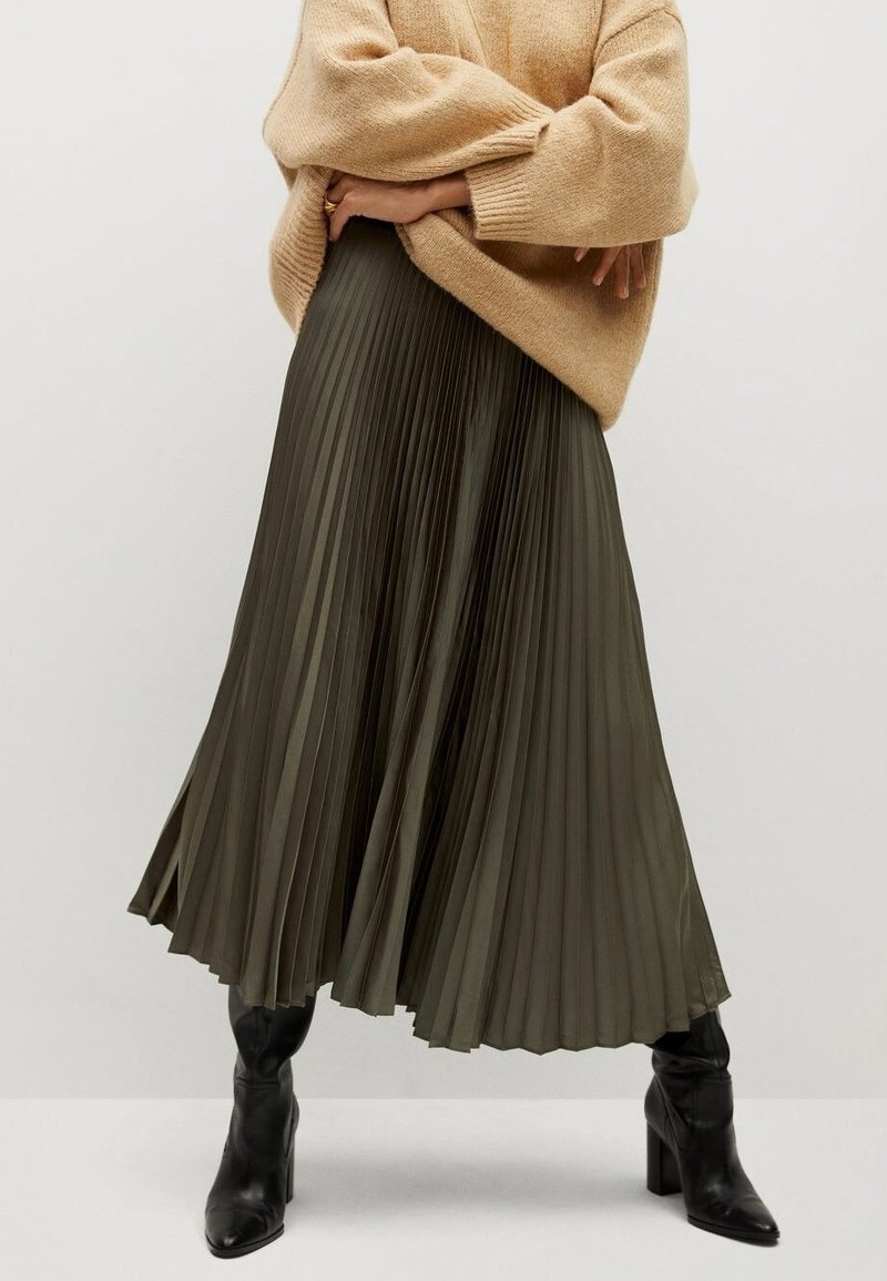 Personne portant un pull beige, une jupe midi plissée vert olive et des bottes à talons noires, debout les bras croisés.