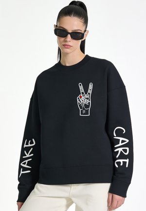 Frau mit dunklem Haar im Pferdeschwanz, die schwarze Sonnenbrillen und einen schwarzen Pullover mit einem Friedenshand-Design und dem Text "Take Care" auf den Ärmeln trägt.