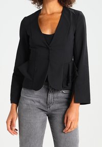 Černý peplum blazer s hlubokým výstřihem do V, dlouhými rukávy a uzávěrem na jeden knoflík; spojený se šedými džíny. Hladká tkanina a přizpůsobený střih.