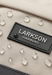 Beige kangasta, jossa on kohotettuja pisteitä, ja musta suorakulmainen etiketti, jossa on valkoisilla kirjaimilla teksti "LARKSON HAMBURG". Teksturoitu pinta, vettä hylkivä.
