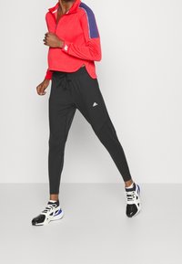 Pullover rosso a maniche lunghe con dettagli blu navy e colletto con zip, abbinato a pantaloni neri affusolati con logo Adidas e vita elasticizzata.