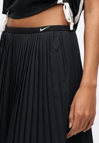 Μαύρη πλισέ φούστα με ελαστική μέση και λευκό λογότυπο Nike. Κατασκευασμένη από μαλακό ύφασμα, που αποκαλύπτει λεπτή υφή και κίνηση.