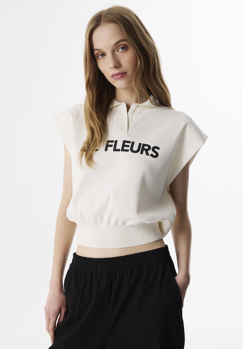 Felpa cropped color crema con colletto polo, maniche corte e lettering nero "FLEURS". Abbinata a pantaloni neri con vita elastica.