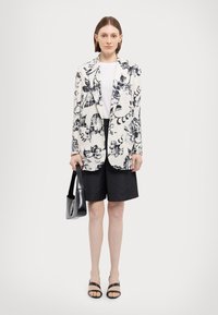 Hvid blazer med navy blomsterprint, parret med sorte shorts og sorte hæle-sandaler. Modellen holder en lille sort taske.