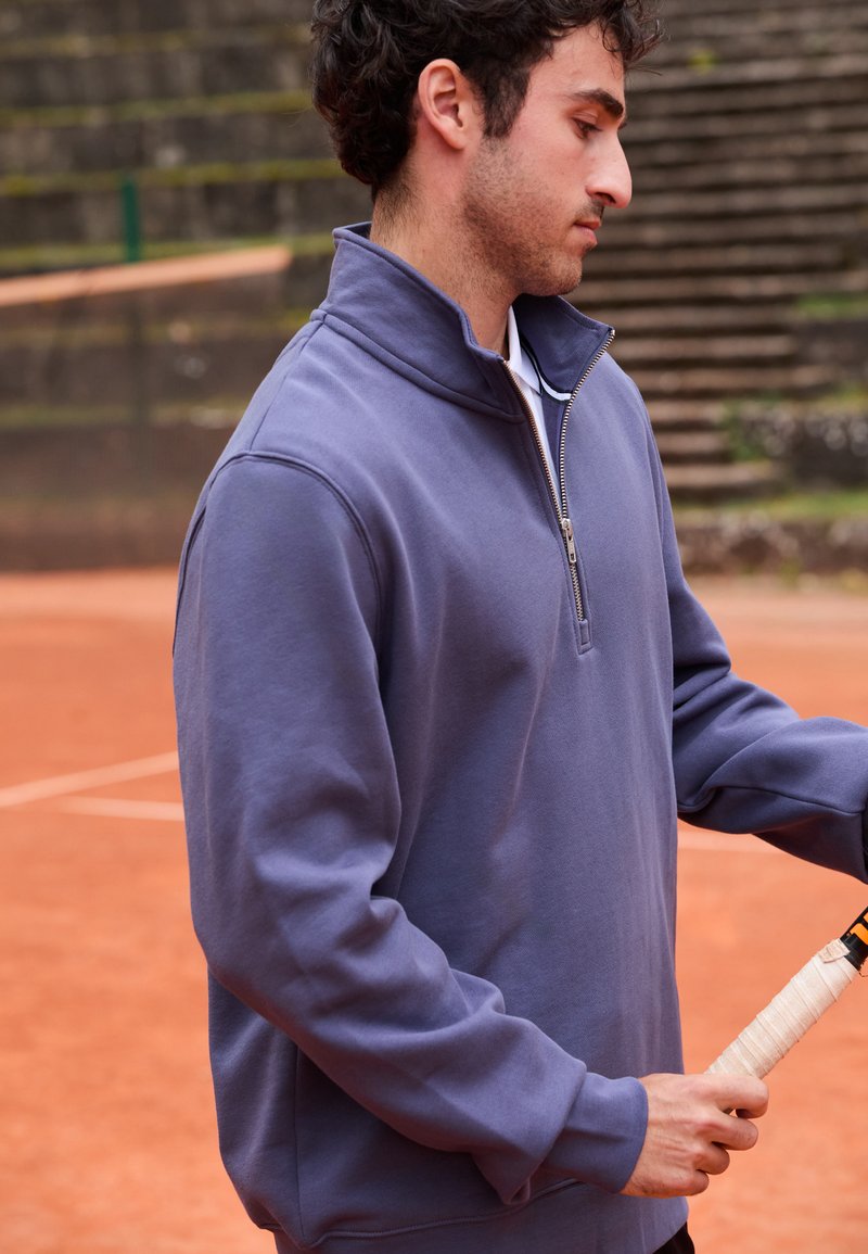 Muž v purpurovom športovom svetri s krátkym zipsom drží tenisovú raketu na antukovom tenisovom kurte s kamennými tribúnami v pozadí.