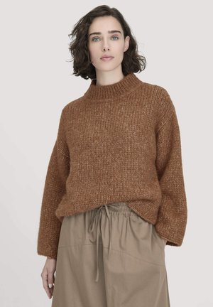 Brauner Strickpullover mit hohem Kragen, lockeren Ärmeln und strukturiertem Finish, kombiniert mit einem hellbeigen, plissierten Rock mit Kordelzug und hohem Bund.