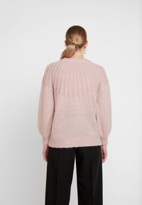 Kobieta z blond włosami w niskim kucyku, ubrana w jasnoróżowy sweter z prążkowanej dzianiny i czarne spodnie z szerokimi nogawkami, zwrócona plecami na białym tle.