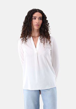 Blusa bianca a maniche lunghe con scollo a V e dettaglio a pieghe, texture liscia e vestibilità ampia, abbinata a jeans azzurri.