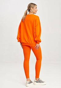 Jasnopomarańczowy, oversize'owy sweatshirt z ściągaczami przy rękawach, w połączeniu z dopasowanymi legginsami oraz białymi sneakersami z czarnym wzorem w panterkę.