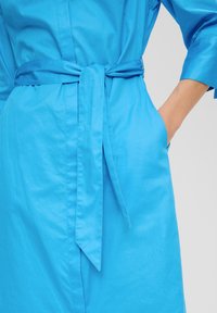 Felle blauwe jurk met een riem in de taille, met een gladde textuur en halflange mouwen. Inclusief zijzakken voor extra functionaliteit.