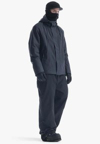 Giacca e pantaloni impermeabili neri, dotati di cappuccio, guanti e una maschera facciale nera. Le calzature sono nere con una superficie testurizzata.