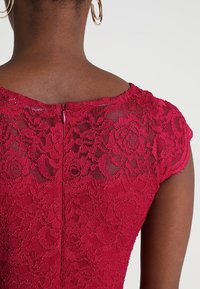 Robe en dentelle bordeaux avec des manches courtes, présentant un motif floral et une fermeture éclair dans le dos. Tissu texturé avec des détails transparents.