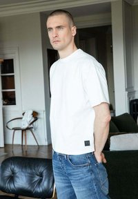 Uomo con capelli corti che indossa una maglietta bianca e jeans blu, in piedi all'interno vicino a mobili in pelle nera e una sedia di legno con un tessuto bianco.