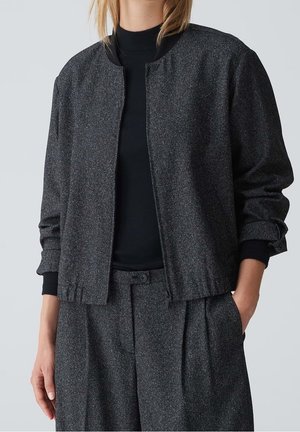 Femme portant une veste bomber texturée gris foncé avec fermeture éclair, un haut noir à manches longues côtelé et un pantalon plissé gris assorti.