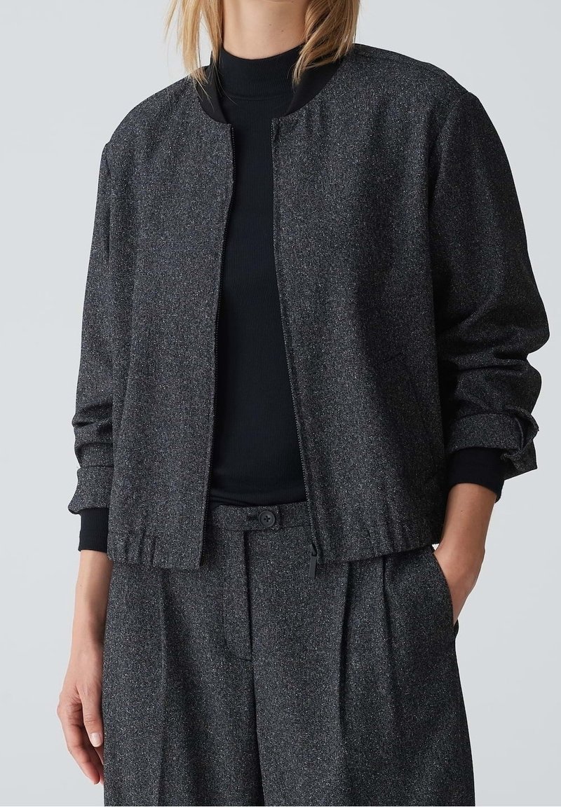 Femme portant une veste bomber texturée gris foncé avec fermeture éclair, un haut noir à manches longues côtelé et un pantalon plissé gris assorti.
