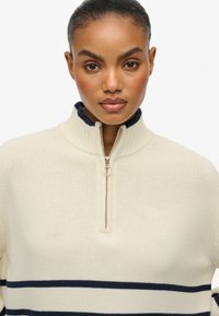 Suéter de color crema con rayas marinas, cuello acanalado y diseño de media cremallera. Presenta un tejido de punto texturizado y un detalle de logo en el pecho.