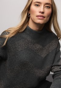 Kobieta o długich blond włosach, nosząca srebrne kolczyki koła oraz ciemnoszary sweter z metalicznymi paskami w kształcie litery V, neutralne tło.