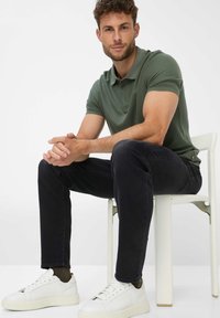 Grünes Poloshirt mit kurzen Ärmeln, dunkle Jeans und weiße Sneaker. Model sitzt auf einem Stuhl, die Hände gefaltet, neutraler Hintergrund.