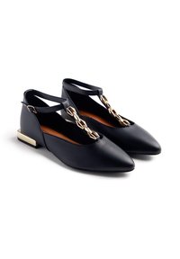 Scarpe flat in pelle nera con punta affilata, cinturino alla caviglia e dettagli in catena dorata, dotate di un piccolo tacco a blocco dorato.