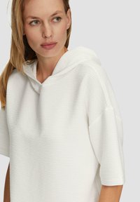 Pull à capuche blanc avec un motif texturé en côtes. Manches courtes et coupe décontractée, avec un large col et un tissu doux.