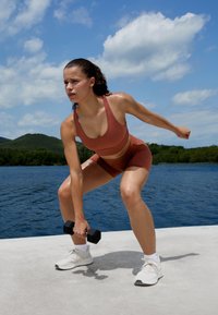 Atletinja v rjavo obarvani športni modrček in hlače, z močnim oprijemom drži črno dumbbell, v ozadju z vodno kuliso in modrim nebom.