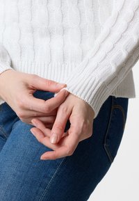 White cable-knit sweater with ribbed cuffs, worn with blue denim jeans. Hands are adjusting the sweater's sleeve. Soft texture visible.