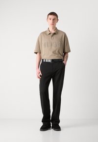Beige poloskjorte med preget "HUGO"-logo, kombinert med svarte bukser og svarte sko. Enkel design, avslappet passform og glatt tekstur.