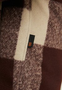 Geruite fleece stof in bruin en crème met een zachte, gestructureerde oppervlakte. Bevat een klein zwart en bruin merklabel.