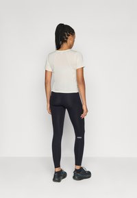 Haut de sport beige clair à manches courtes et leggings noirs avec logo. Les leggings sont ajustés, fabriqués dans un matériau extensible et présentent un design longueur cheville.