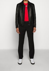 Person iført en sort læderjakke, rød skjorte, sorte bukser og hvide sneakers, der står imod en ensfarvet lys baggrund.