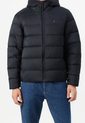 Person trägt eine schwarze gesteppte Daunenjacke mit Reißverschluss vorne und Kapuze, kombiniert mit blauen Jeans vor schlichtem Hintergrund.