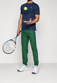 Man med marinblå skjorta och gröna träningsbyxor som håller en tennisracket och kastar en tennisboll framför en enfärgad bakgrund.