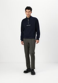 Marineblå zip-up sweatshirt med kengurulomme og broderet logo, parret med grå plaid skræddersyede bukser og sorte sko.