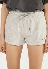 Frau trägt hellgraue Fleece-Shorts mit elastischem Kordelzugbund und kleinem "ESCAPE"-Schriftzug am unteren rechten Saum.