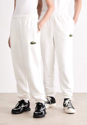 Zwei Personen tragen weiße Jogginghosen mit grünen Krokodil-Logos und schwarze Sneakers, stehen nebeneinander mit den Händen in den Taschen.