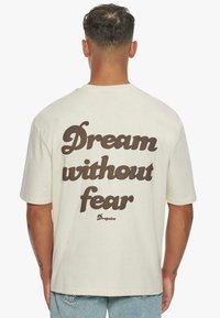 Cremefarvet bomulds t-shirt, korte ærmer, med fed brun tekst "Drøm uden frygt" på ryggen, lige snit, afslappet design.