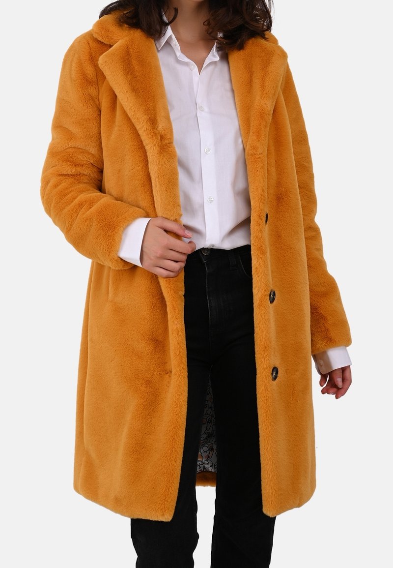 Sinapinkeltainen tekoturkistakki, jossa on kauluskäänteet, yksittäinen nappisuljenta ja kaksi sivutaskua, pidetty valkoisen paidan ja mustien farkkujen päällä.