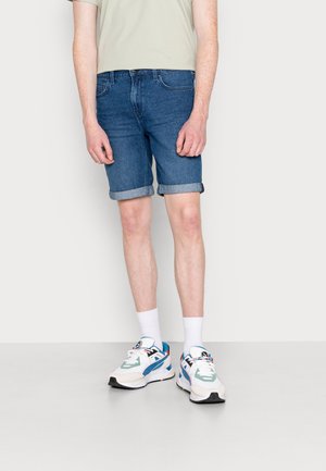 Man draagt opgerolde blauwe spijkershorts, witte sokken en witte sneakers met blauwe en groene accenten, staand tegen een effen achtergrond.