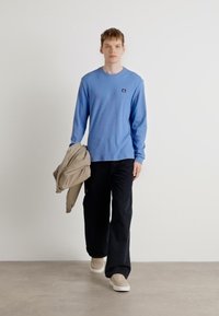 Blå stripet longsleeve skjorte, mørke vide bukser, beige jakke over den ene armen, og beige sneakers, stående mot en nøytral bakgrunn.