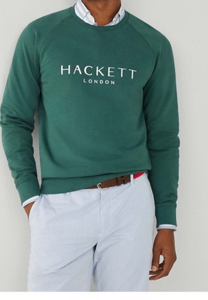 Personne portant un sweat-shirt vert Hackett London par-dessus une chemise blanche à col rentrée dans un pantalon bleu clair rayé avec une ceinture marron.