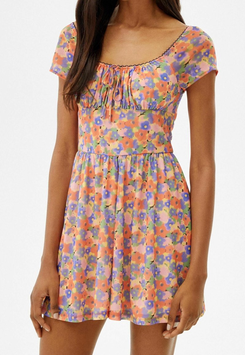 Femme portant une mini-robe à manches courtes fleurie avec des fleurs orange, violettes et roses, avec un col froncé et un nœud devant.