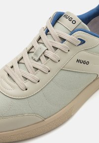Baskets vert clair avec un upper texturé, lacets beiges et accents bleus. Présente un logo sur le côté et une semelle en caoutchouc.