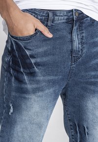 Vaqueros de denim en lavado medio, con un ligero desvanecimiento y detalles desgastados. Diseño clásico de cinco bolsillos con cierre de botón y cremallera.