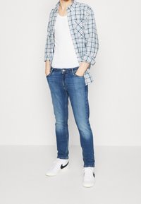 Modrá kockovaná košile přes bílou tílko, doplněná slim-fit modrými džínami a bílými teniskami. Košile má přední kapsy.